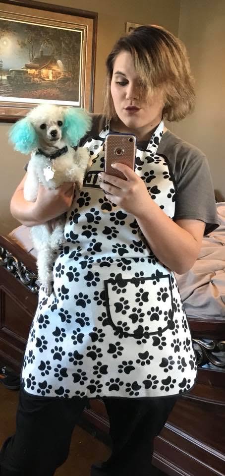 Woman wearing a paw print apron takes a mirror selfie while holding a white poodle with teal-dyed ears