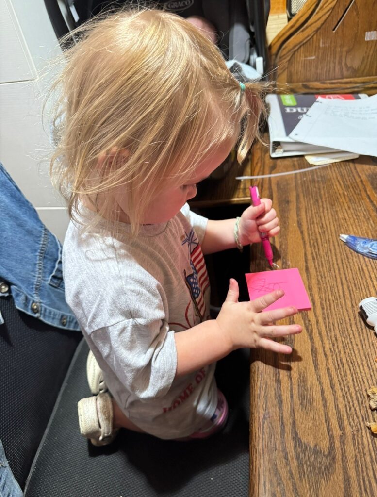 Toddler drawing on a pink sticky note with a marker at a wooden table